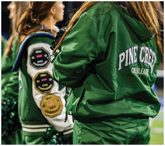 People wearing green cheerleading jackets with “Pine Crest Cheerleader" on the back stand in a row, some holding green pom-poms. Description generated by AI