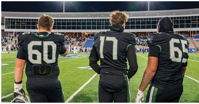 Three people wearing black football uniforms with numbers 60, 17, and 69 stand on a football field facing away from the camera. Description generated by AI