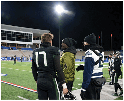 Three people in sports attire are standing and talking on a football field at night with a stadium and other players in the background. Description generated by AI