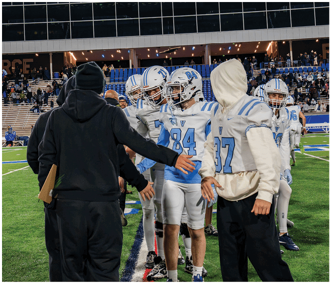 People in football uniforms and hoodies are gathered on a football field, appearing to be in discussion or preparing for a game. Description generated by AI