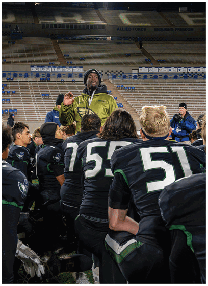 A group of people in black sports uniforms with numbers kneel on a field while one person in a green jacket stands and speaks to them in a stadium with empty bleachers. Description generated by AI