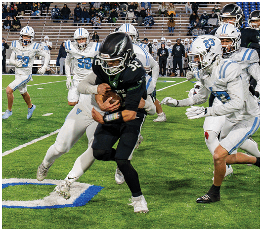 People in football uniforms are actively playing on a field with one person in black running with the ball while others in white attempt to tackle or block. Description generated by AI