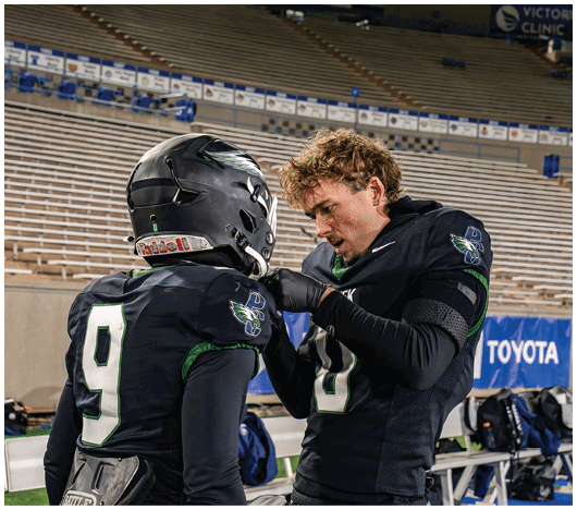Two people in football uniforms are engaged in an intense conversation on the sidelines of a stadium with empty bleachers in the background. Description generated by AI