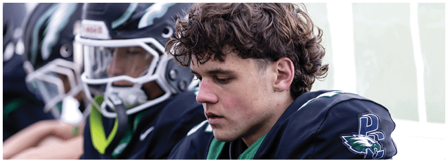 A person with curly hair wearing a dark sports uniform is sitting next to others in similar helmets and uniforms, appearing focused or contemplative. Description generated by AI