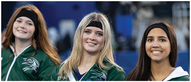 Three smiling adults wearing black headbands and green outfits stand closely together in an outdoor setting. Description generated by AI