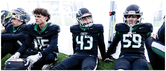 Four people in football uniforms and helmets are sitting on a bench inside a tent, appearing to rest or wait during a game or practice. Description generated by AI