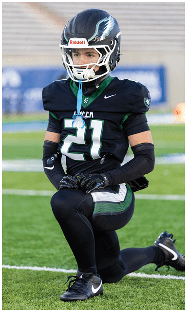 A person wearing a black football uniform with the number 21 and a helmet is kneeling on one knee on a football field. Description generated by AI