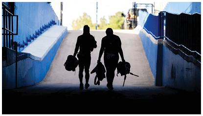 Two people carrying helmets and gear walk up a ramp in a tunnel with blue walls toward a bright outdoor area. Description generated by AI