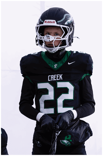 A person wearing a black football uniform with the number 22 and the word Creek on the jersey is standing while holding a black bag and wearing a helmet with a faceguard. Description generated by AI