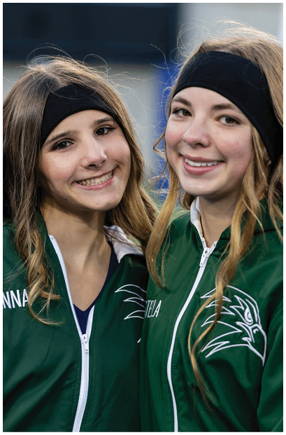 A person with long hair wearing a green jacket with the name “JENNA" embroidered on it and a black headband is smiling outdoors. Description generated by AI