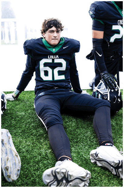 A person wearing a black and green football uniform with the number 62 is sitting on artificial turf with legs extended and a focused expression. Description generated by AI