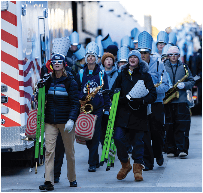 A group of people wearing matching tall hats and casual winter clothing are walking outdoors, some carrying musical instruments and equipment. Description generated by AI