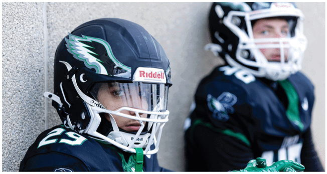Two people wearing football helmets and uniforms are sitting against a wall, with one looking directly at the camera and the other looking away. Description generated by AI