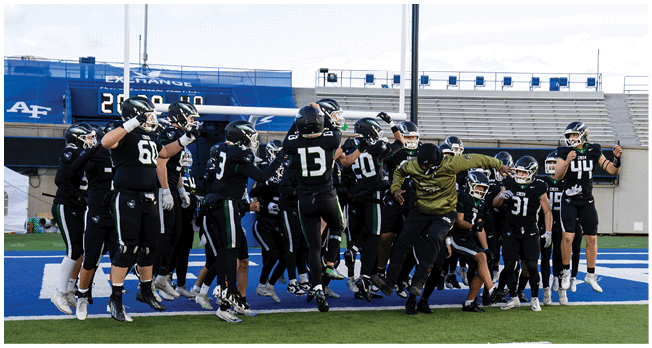 A group of people wearing black sports uniforms and helmets are gathered on a field, some raising their arms in celebration or excitement. Description generated by AI