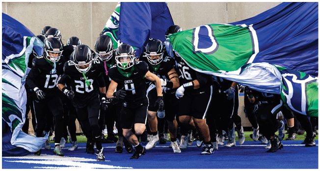 A group of people in black sports uniforms and helmets run together through large blue and green flags in a stadium. Description generated by AI
