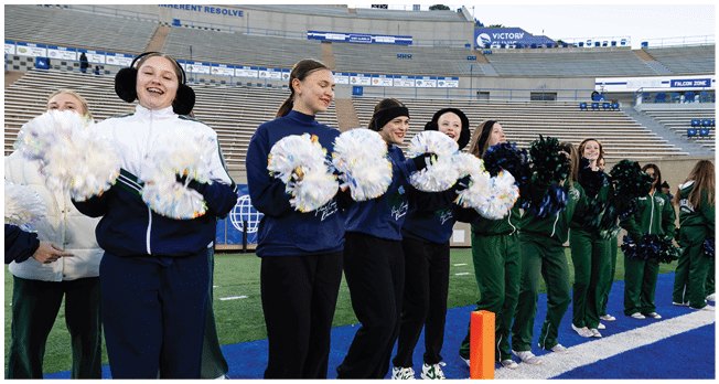 A group of people dressed in coordinated outfits are standing in a line on a sports field holding pom-poms, with empty stadium seats in the background. Description generated by AI