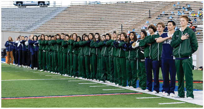 A large group of people dressed in green and blue athletic uniforms stand in a line on a sports field with their hands over their hearts, facing forward. Description generated by AI