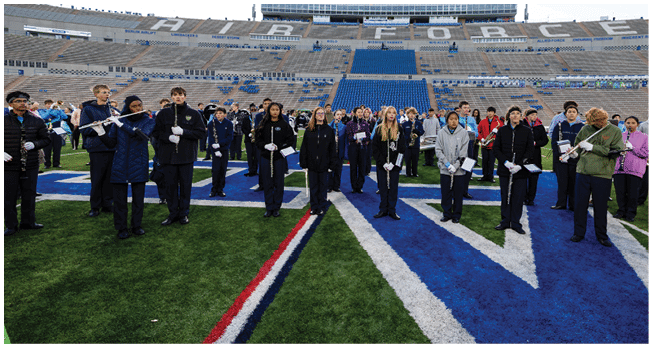 A large group of people dressed in dark clothing are standing on a football field holding musical instruments, arranged in rows with a stadium in the background. Description generated by AI