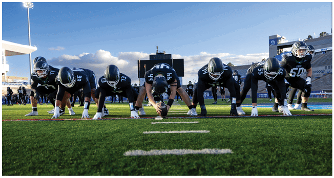 Four people in football uniforms are lined up on a field in a ready stance under a clear sky. Description generated by AI