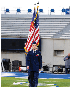 A person in a formal military uniform is walking on a sports field carrying two flags, one of which is the United States flag, with empty bleachers in the background. Description generated by AI