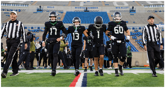 A referee and two football players in black uniforms with helmets walk on a football field with empty blue stadium seats in the background. Description generated by AI