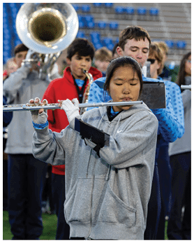 A person wearing a gray hoodie and white gloves is playing a flute in a marching band with other people playing various instruments in a stadium setting. Description generated by AI