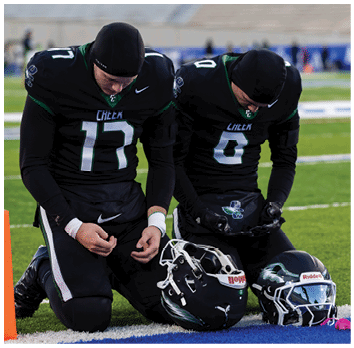 Two people in black football uniforms kneel on a field with their helmets placed on the ground in front of them, appearing to be in a moment of reflection or prayer. Description generated by AI