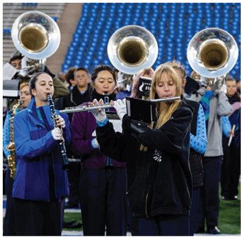 People wearing jackets and gloves are playing various wind instruments in a marching band on a sports field with blue stadium seats in the background. Description generated by AI