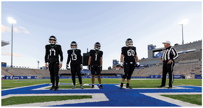 Four people in black football uniforms stand on a football field with a referee nearby and empty stadium seats in the background. Description generated by AI