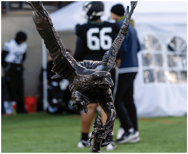 A person wearing a black sports jersey with the number 66 stands next to another person in a dark jacket and hat behind a large sculpture with tall, curved antlers. Description generated by AI