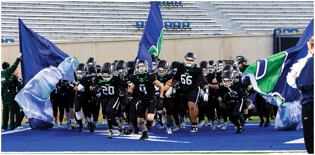 A group of football players in black uniforms with helmets are running onto a blue field through large flags being held up on either side. Description generated by AI
