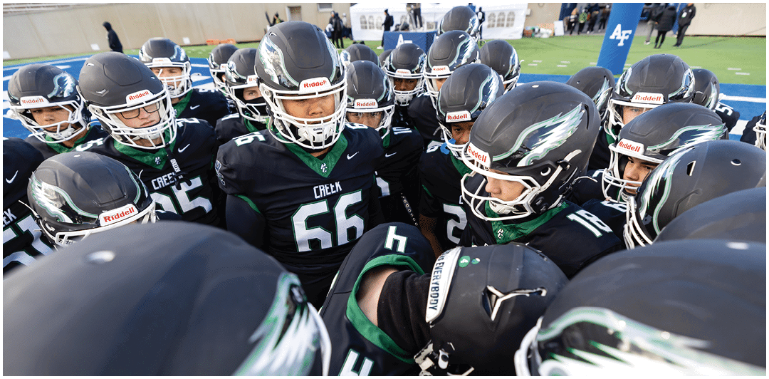 A group of football players wearing black uniforms and helmets are huddled together on a field inside a stadium. Description generated by AI