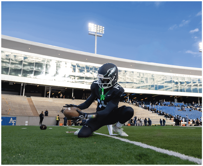 A person in a dark football uniform is kneeling on a field while holding a football near a white line in a stadium with empty and partially filled bleachers under a clear sky. Description generated by AI