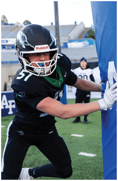 A person wearing a black football uniform and helmet is pushing against a blue padded blocking sled on a football field. Description generated by AI