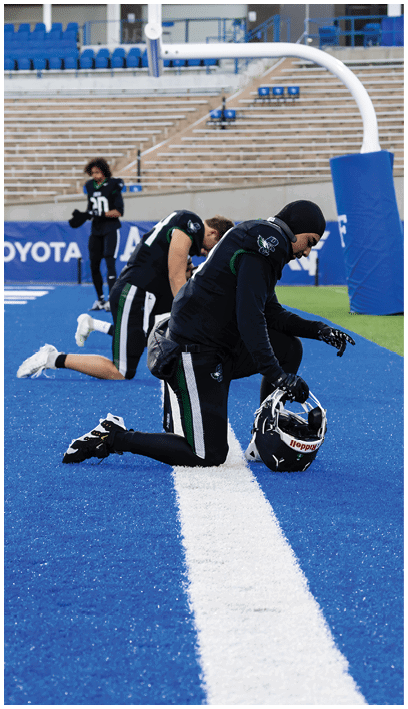 People in sports uniforms are kneeling and standing on a blue football field near the end zone with empty stadium seats in the background. Description generated by AI