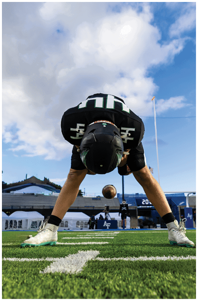 An athlete wearing a black uniform and cleats is in a crouched starting position on a green football field with a stadium and cloudy sky in the background. Description generated by AI