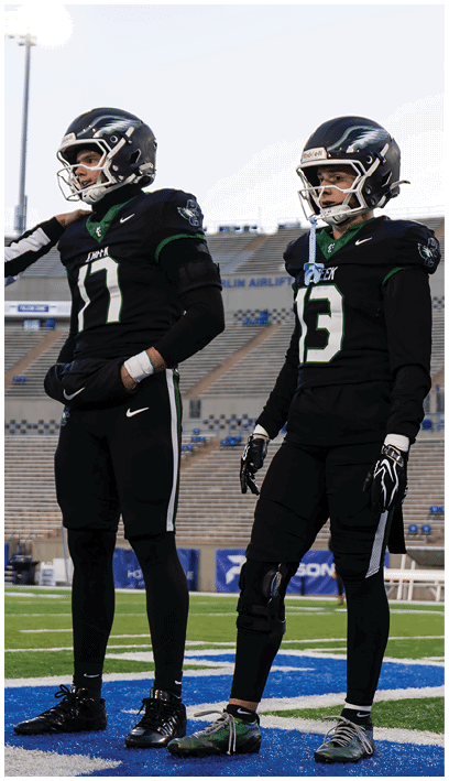 Two people wearing black football uniforms and helmets stand on a football field with empty stadium seats in the background. Description generated by AI