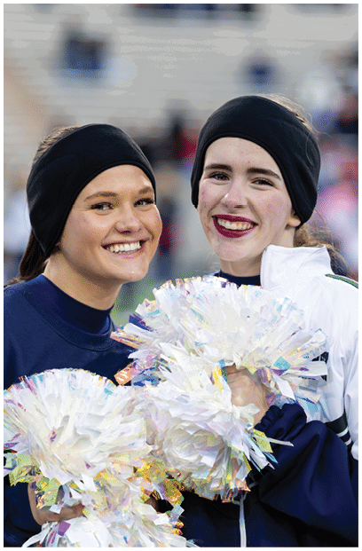 A person wearing a dark headband and sweatshirt is smiling and holding iridescent pom-poms on a sports field with blurred officials in the background. Description generated by AI