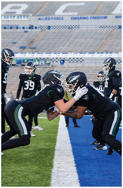 Two people in football uniforms and helmets are facing each other closely in a stadium with empty bleachers in the background while another person stands nearby. Description generated by AI