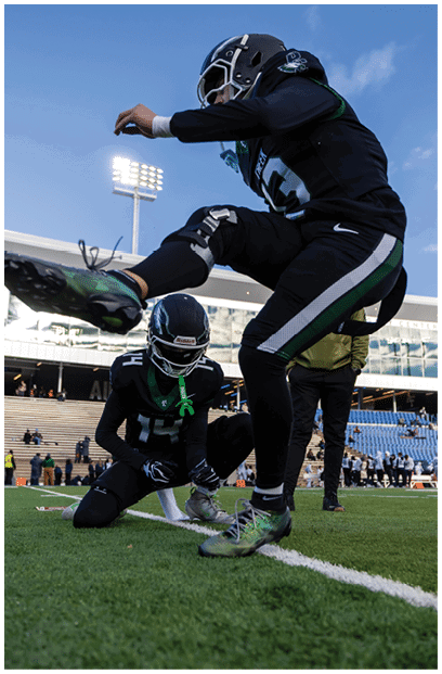 Two people in football uniforms are on a field with one person kneeling and the other mid-jump during a game or practice in a stadium. Description generated by AI