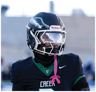 A person wearing a black football uniform with green accents and a helmet with a clear visor is on a field, smiling. Description generated by AI