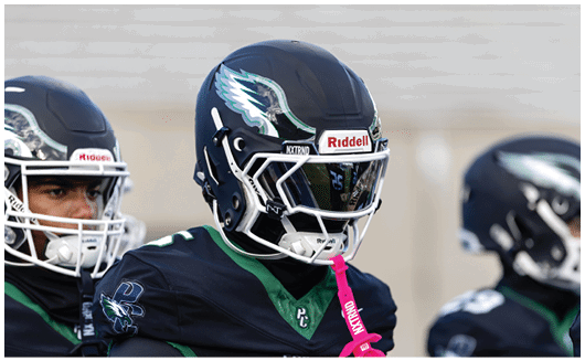 Three people wearing dark football uniforms and helmets with an eagle logo are standing on a field. Description generated by AI