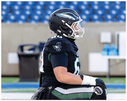 A person wearing a black football uniform and helmet is sitting on the sidelines of a stadium with empty blue seats in the background. Description generated by AI