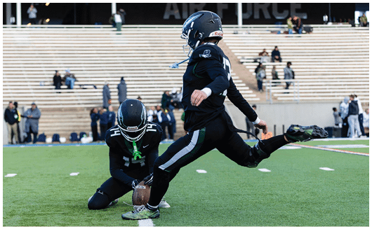 Two people in black football uniforms are on a field with one holding the ball on the ground while the other prepares to kick it. Description generated by AI