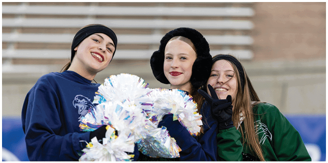 Three people wearing winter clothing and smiling hold shiny pom-poms in an outdoor setting with bleachers in the background. Description generated by AI