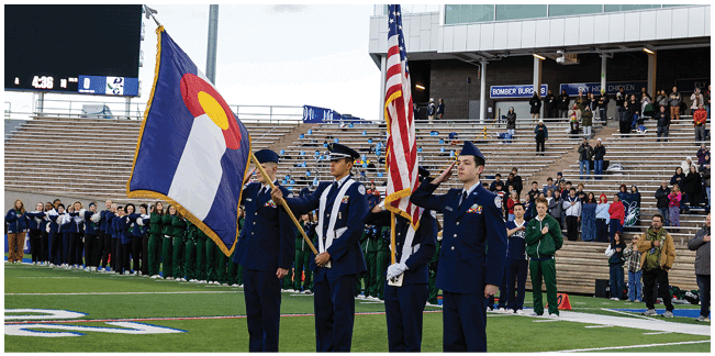 People in uniform hold flags while standing in front of a crowd seated on stadium bleachers during an outdoor event. Description generated by AI