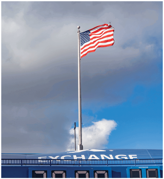 An American flag waves on a tall flagpole above a blue structure with the word “EXCHANGE" partially visible against a cloudy sky. Description generated by AI