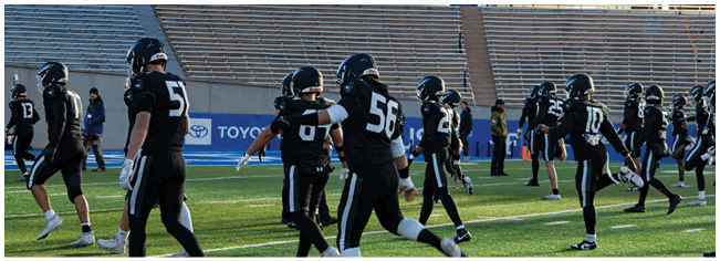 A group of people wearing black sports uniforms and helmets are gathered on a field with empty bleachers in the background. Description generated by AI
