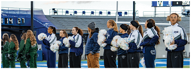 A group of people in matching uniforms stand in a line on a sports field with empty bleachers and a scoreboard in the background. Description generated by AI