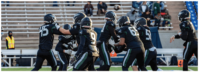 A group of football players in dark uniforms are engaged in a play on the field with empty bleachers and scattered spectators in the background. Description generated by AI